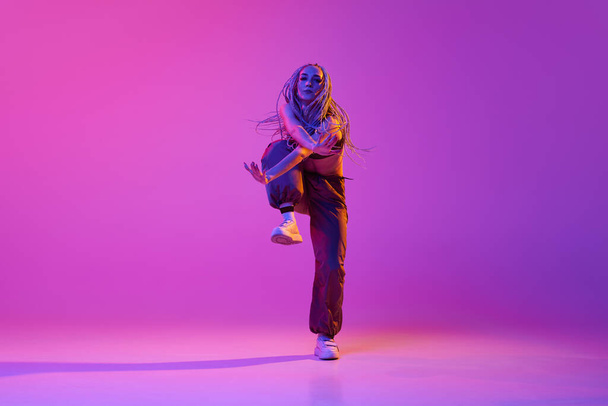 Romper baile al estilo de la calle. Una hermosa chica, joven bailarina moderna haciendo trucos de hip-hop sobre fondo de estudio púrpura en luz de neón. Concepto de estilo contemporáneo, movimiento, arte, movimiento, inspiración, anuncio - Foto, imagen