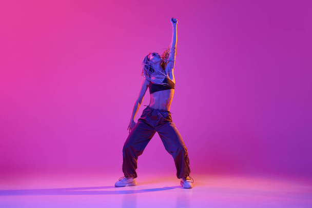 Jonge mooie slanke meisje, moderne danseres dragen hip-hop kleding dansen over gradiënt paars neon achtergrond. Concept van hedendaagse dansstijl, inspiratie, beweging, beweging, hobby, mode, kunst - Foto, afbeelding