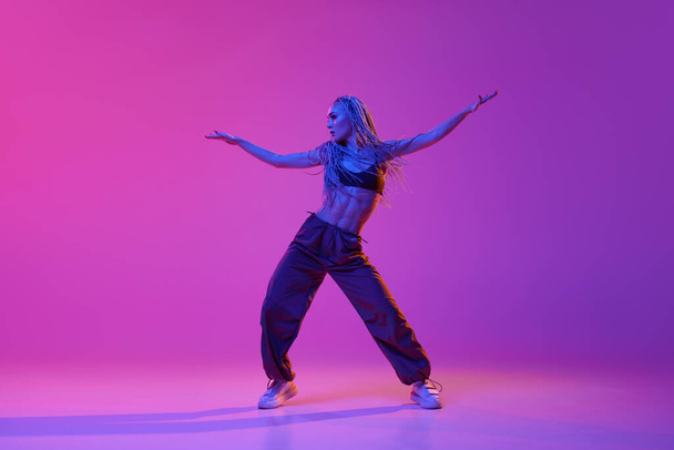 Freedom. Stylish sporty girl, modern dancer moving to the beat of music over purple studio background in neon light. Concept of contemporary dance style, motion, art, youth, culture, music, hobby, ad - Photo, Image