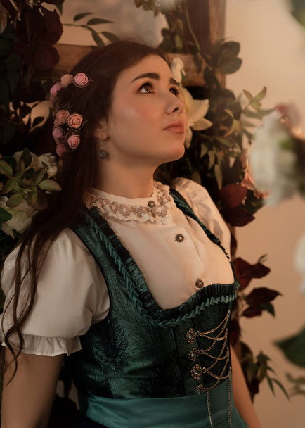 A young peasant woman standing in a garden with flowers dressed in a 19th century bavarian dress. - Foto, afbeelding