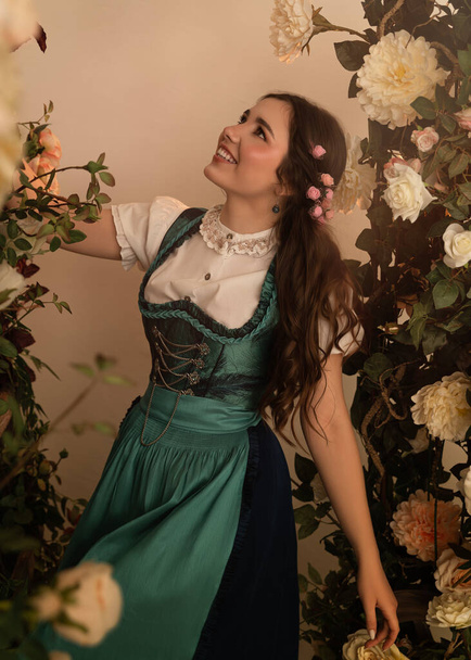 A young peasant woman standing in a garden with flowers dressed in a 19th century bavarian dress. - Foto, afbeelding