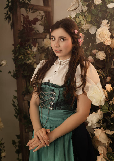 A young peasant woman standing in a garden with flowers dressed in a 19th century bavarian dress. - Фото, изображение
