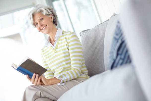 Settling down with a good book. Portrait of a happy senior woman reading a book on the sofa in her home - Foto, Imagen