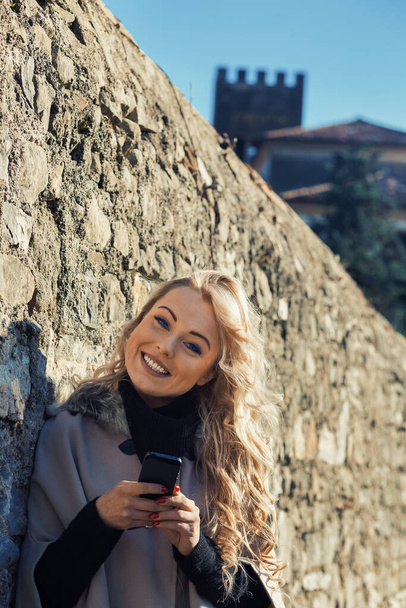Young woman with long, curly, perfectly permed blonde hair, perfect makeup and manicure, is a tourist in a European city with medieval villages. Using her phone to talk, chat, or access Wi-Fi for hist - Photo, Image