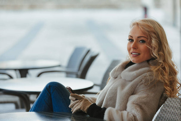 Όμορφη νεαρή ξανθιά γυναίκα κάθεται σε ένα μπαρ, διασκεδάζοντας, γελώντας με κάτι ξεκαρδιστικό στο smartphone της ή ένα ληφθέν μήνυμα. Είναι τουρίστρια σε μια ευρωπαϊκή πόλη, απολαμβάνοντας τον καθαρό αέρα, ντυμένη  - Φωτογραφία, εικόνα