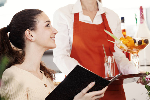 Mooie brunette jonge vrouw in de koffiebar, ijssalon of banketbakker met een zwart menu op haar handen glimlacht terwijl ze een kopje fruitsalade met ijs op de top bestelt bij de ijssalon. - Foto, afbeelding