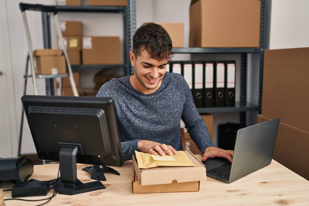 Young hispanic man ecommerce business worker using laptop holding packages at office - Фото, зображення