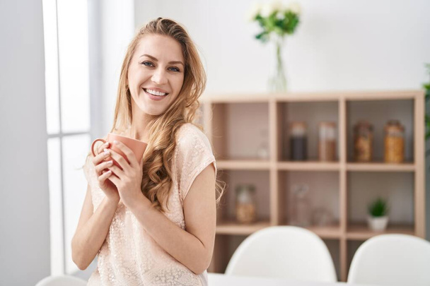 Νεαρή ξανθιά γυναίκα χαμογελά αυτοπεποίθηση πίνοντας καφέ στο σπίτι - Φωτογραφία, εικόνα