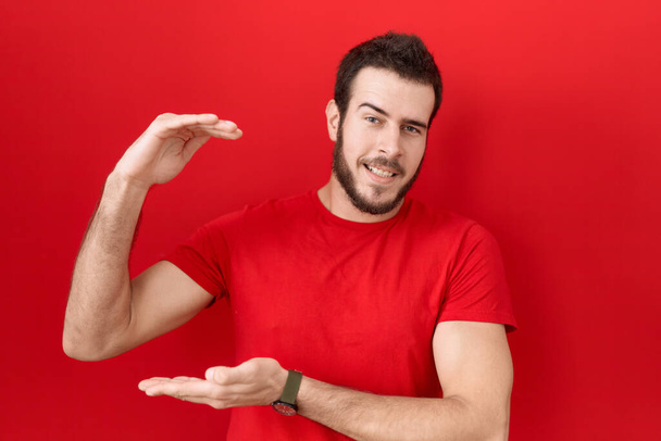 Молодий іспаномовний чоловік у повсякденній червоній сорочці жестикулює руками, показуючи великий і великий знак розміру, мірний символ. усміхнений погляд на камеру. концепція вимірювання
.  - Фото, зображення