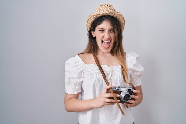 Young brunette woman holding vintage camera sticking tongue out happy with funny expression.  - Foto, afbeelding