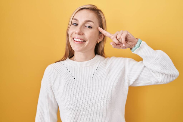 Young caucasian woman wearing white sweater over yellow background smiling pointing to head with one finger, great idea or thought, good memory  - Fotó, kép