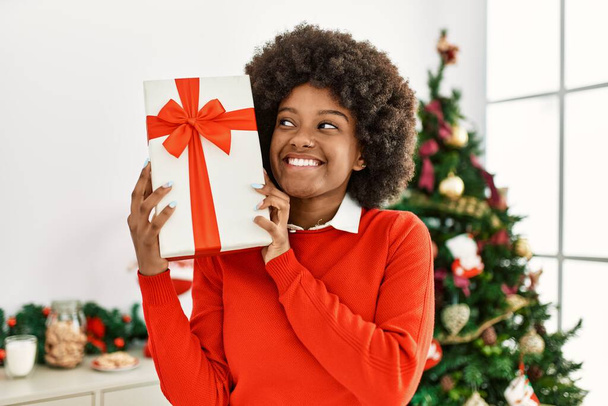 Young african american woman holding gift standing by christmas tree at home - Fotoğraf, Görsel