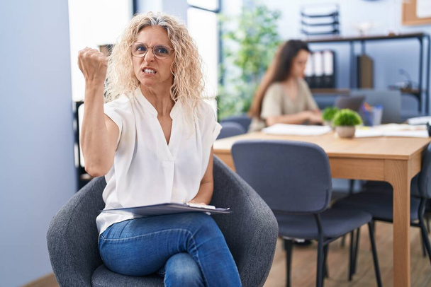 Kobieta w średnim wieku pracująca w biurze trzymająca notatnik zirytowana i sfrustrowana krzykiem gniewu, wrzeszcząca szaleństwem ze złości i podniesionymi rękami  - Zdjęcie, obraz