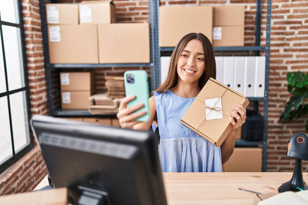 Young beautiful hispanic woman ecommerce business worker holding package make selfie by smartphone at office - 写真・画像