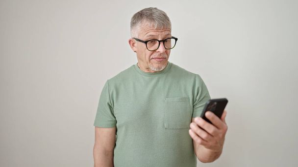 Grauhaariger Mann mittleren Alters benutzt Smartphone mit ernstem Gesichtsausdruck vor isoliertem weißem Hintergrund - Foto, Bild