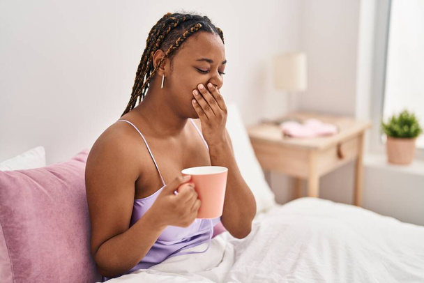 Femme afro-américaine assise sur le lit bâillant et buvant du café dans la chambre - Photo, image