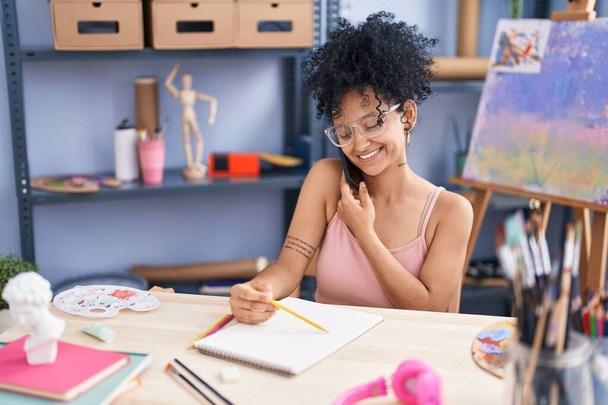 Молодая латиноамериканка-художница разговаривает на смартфоне, рисуя на ноутбуке в арт-студии - Фото, изображение