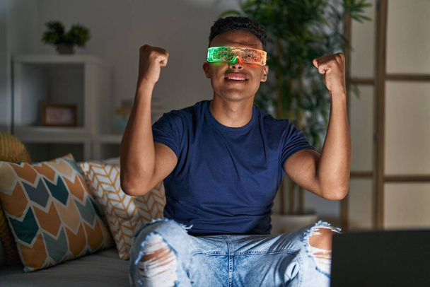 Mladý latin muž pomocí virtuální realita brýle sedí na pohovce doma - Fotografie, Obrázek
