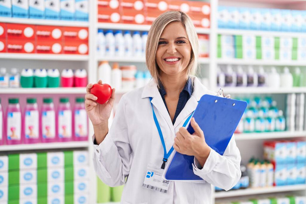 Jovem mulher trabalhando na farmácia farmácia segurando o coração sorrindo com um sorriso feliz e legal no rosto. mostrando dentes.  - Foto, Imagem