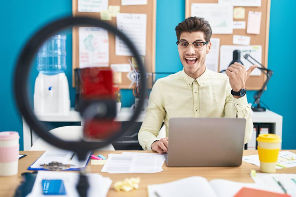 Nuori latinomies tekee videopuhelun älypuhelimella toimistossa osoittaen peukaloa sivulle hymyillen onnellisena avoimella suulla  - Valokuva, kuva