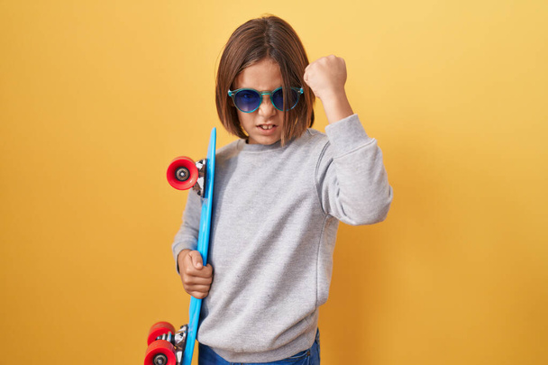 Kleiner hispanischer Junge mit Sonnenbrille und Skateboard, genervt und frustriert schreiend vor Wut, schreiend vor Wut und erhobener Hand  - Foto, Bild