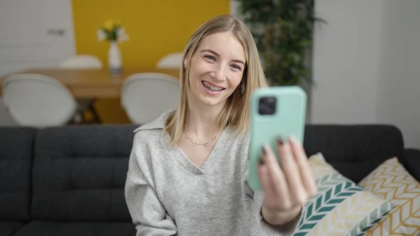 Young blonde woman taking selfie picture with smartphone sitting on the sofa at home - Фото, изображение