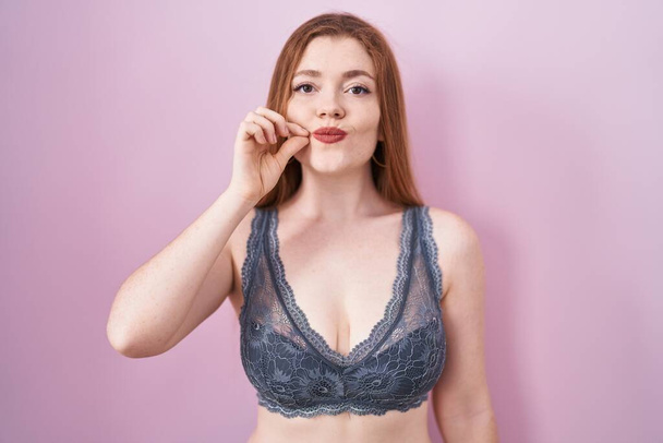 Redhead woman wearing lingerie over pink background mouth and lips shut as zip with fingers. secret and silent, taboo talking  - Фото, изображение