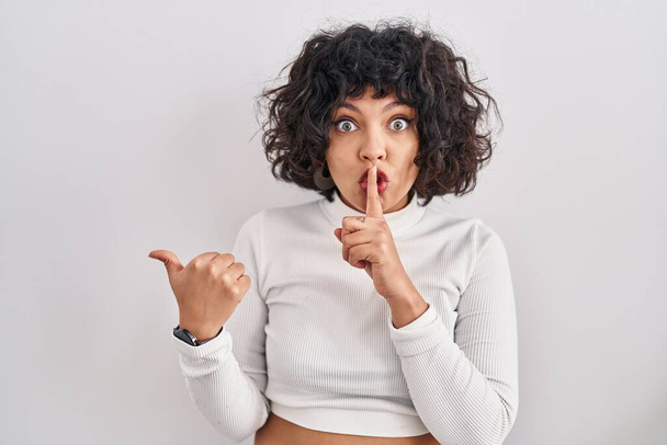 Hispanic woman with curly hair standing over isolated background asking to be quiet with finger on lips pointing with hand to the side. silence and secret concept.  - Photo, Image