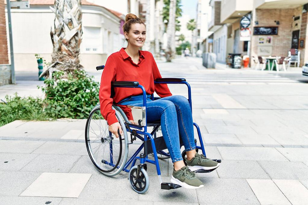Jonge vrouw glimlachend zelfverzekerd zittend op rolstoel op straat - Foto, afbeelding