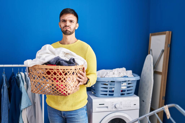 Hispanic man holding laundry basket puffing cheeks with funny face. mouth inflated with air, catching air.  - Fotoğraf, Görsel