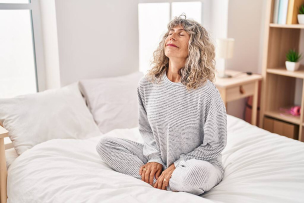 Жінка середнього віку займається вправами йоги, сидячи на ліжку в спальні
 - Фото, зображення