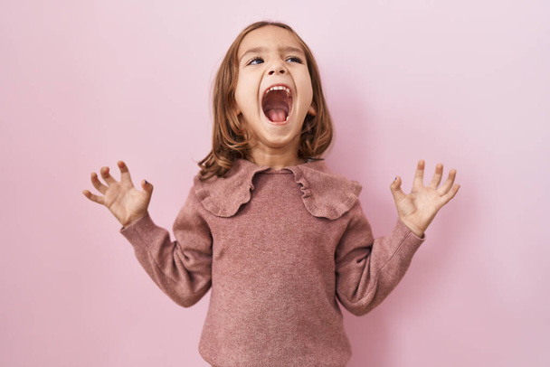Little hispanic girl standing over pink background crazy and mad shouting and yelling with aggressive expression and arms raised. frustration concept.  - Foto, Imagem