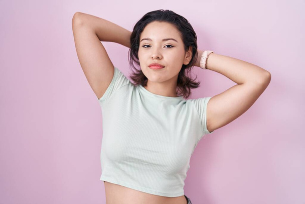 Hispanic young woman standing over pink background relaxing and stretching, arms and hands behind head and neck smiling happy  - Foto, Imagem