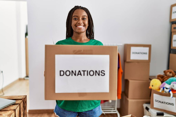 Young african american woman wearing volunteer uniform holding donations package at charity center - Φωτογραφία, εικόνα