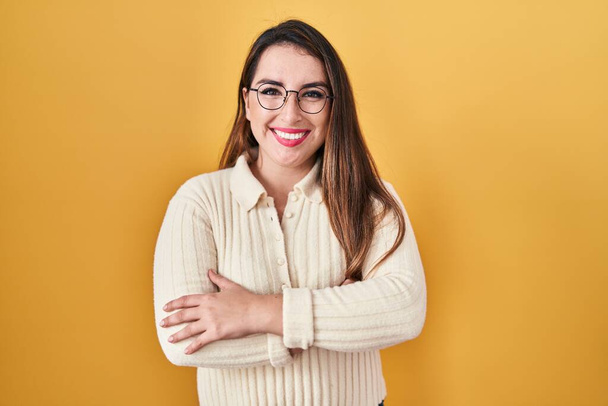 Young hispanic woman standing over yellow background happy face smiling with crossed arms looking at the camera. positive person.  - Foto, imagen