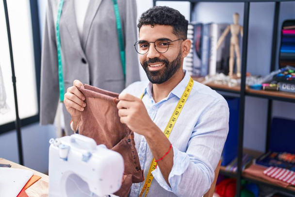 Junger arabischer Schneider lächelt selbstbewusst und hält Jeans in der Schneiderei - Foto, Bild