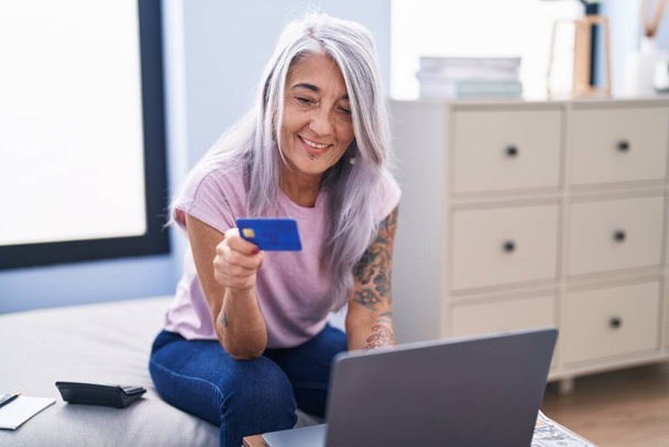 Middelbare leeftijd grijs-harige vrouw met behulp van laptop en creditcard zittend op de bank thuis - Foto, afbeelding