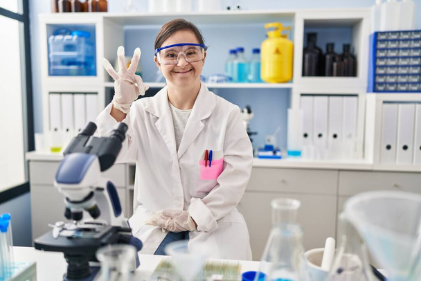 Chica hispana con síndrome de Down que trabaja en el laboratorio científico sonriendo positiva haciendo signo de ok con la mano y los dedos. expresión exitosa.  - Foto, imagen