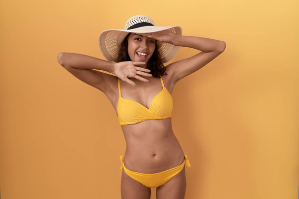 Jonge Spaanse vrouw in bikini en zomerhoed lachend vrolijk kiekeboe spelend met wijzers met gezicht. verrast en verlaten  - Foto, afbeelding