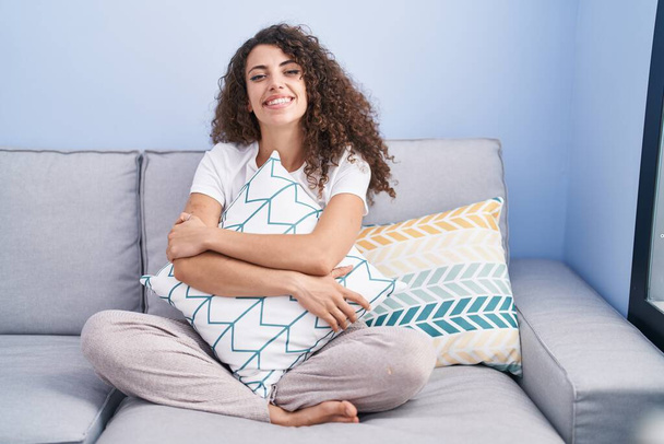 Hispanische Frau mit lockigem Haar, die zu Hause auf dem Sofa sitzt und mit einem fröhlichen und kühlen Lächeln im Gesicht lächelt. Zähne zeigen.  - Foto, Bild
