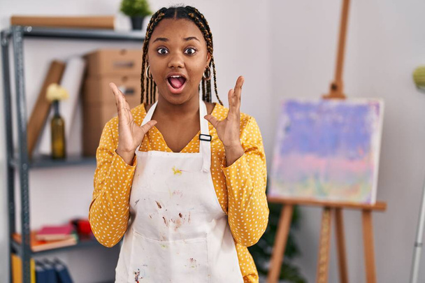 Africká americká žena s copánky v uměleckém studiu slaví vítězství se šťastným úsměvem a vítězným výrazem se zdviženýma rukama  - Fotografie, Obrázek