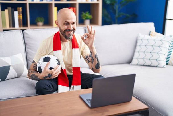 Hispanischer Mann mit Tätowierungen beobachtet Fußballspiel Hooligan hält Ball auf dem Laptop tut ok Zeichen mit den Fingern, lächelt freundlich gestikulierend hervorragendes Symbol  - Foto, Bild