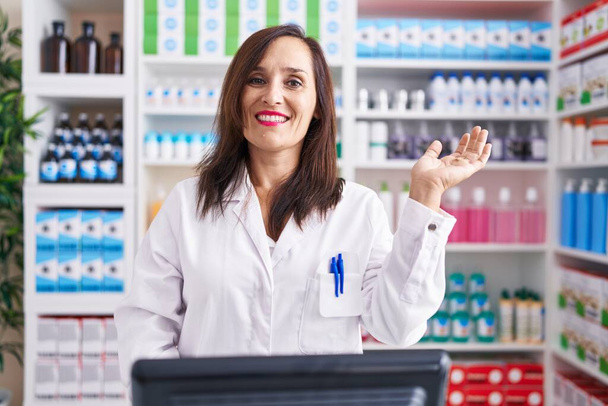 Middle age brunette woman working at pharmacy drugstore smiling cheerful presenting and pointing with palm of hand looking at the camera.  - Photo, Image