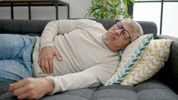 Hombre de pelo gris de mediana edad que sufre de dolor de estómago acostado en el sofá en casa - Foto, Imagen