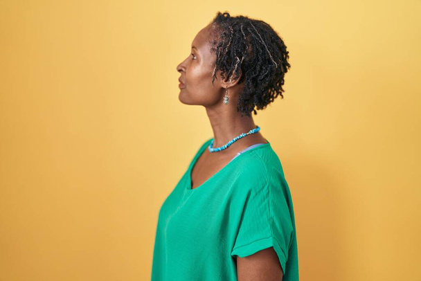 Αφρικανική γυναίκα με dreadlocks στέκεται πάνω από κίτρινο φόντο κοιτάζοντας προς τα πλάγια, να χαλαρώσετε προφίλ ποζάρουν με φυσικό πρόσωπο και αυτοπεποίθηση χαμόγελο.  - Φωτογραφία, εικόνα