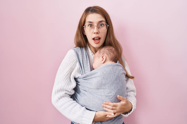 Nuori valkoihoinen nainen tilalla ja kuljettaa vauvaa rintareppu peloissaan ja hämmästynyt suu auki yllätys, epäusko kasvot  - Valokuva, kuva