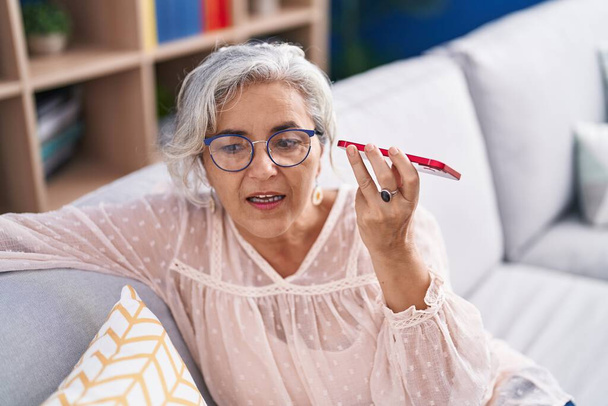 Keski-ikäinen harmaa tukka nainen kuuntelee ääniviestin älypuhelimella istuu sohvalla kotona - Valokuva, kuva
