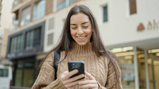 Νεαρή όμορφη ισπανόφωνη γυναίκα που χρησιμοποιεί smartphone χαμογελώντας στο δρόμο - Φωτογραφία, εικόνα
