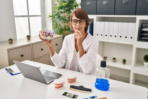 Brunette dentist woman holding sweets serious face thinking about question with hand on chin, thoughtful about confusing idea  - Photo, Image