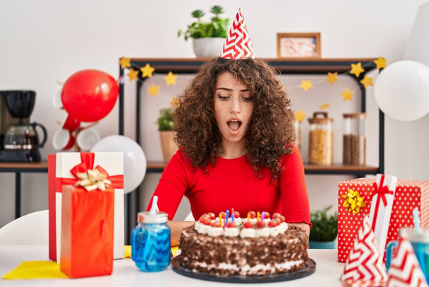 Hispanic woman with curly hair celebrating birthday holding big chocolate cake scared and amazed with open mouth for surprise, disbelief face  - Photo, Image